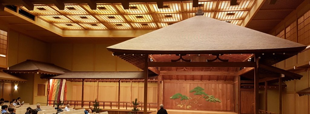 A simple roof sits over a nogaku stage under the ceiling lights of the larger building in which the whole stage is enclosed