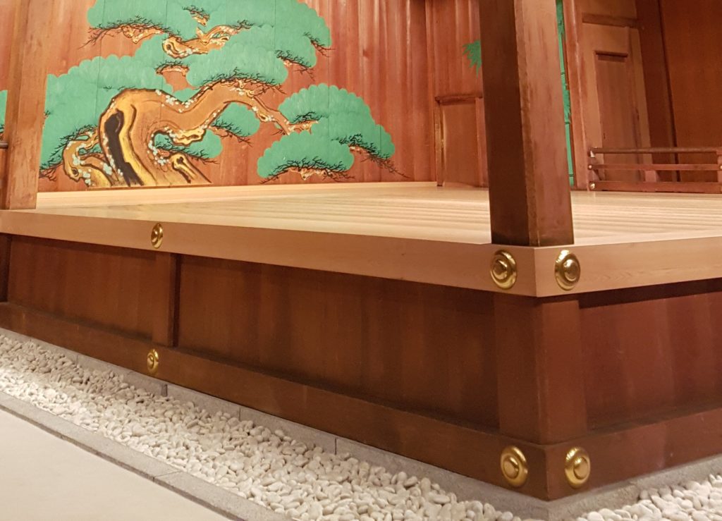 A boarder of white rocks cover the ground surrounding the base of the noh stage 