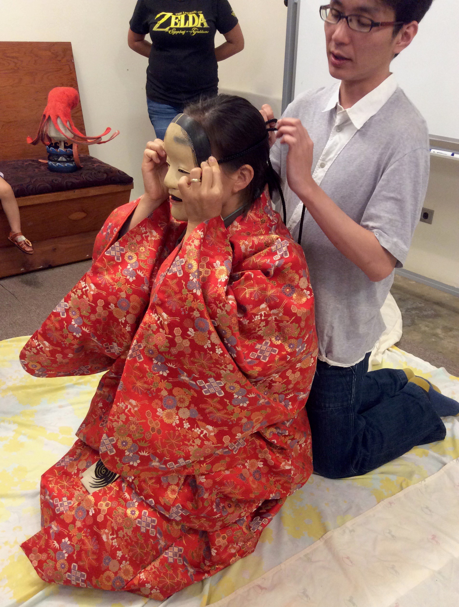 Shigeyama Sennojō III ties a noh mask onto a student in a red brocade kimono