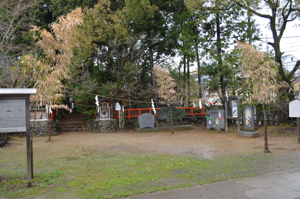 An information sign, stone monuments and trees occupy the perimeter of an open square at Imagumano Shrine