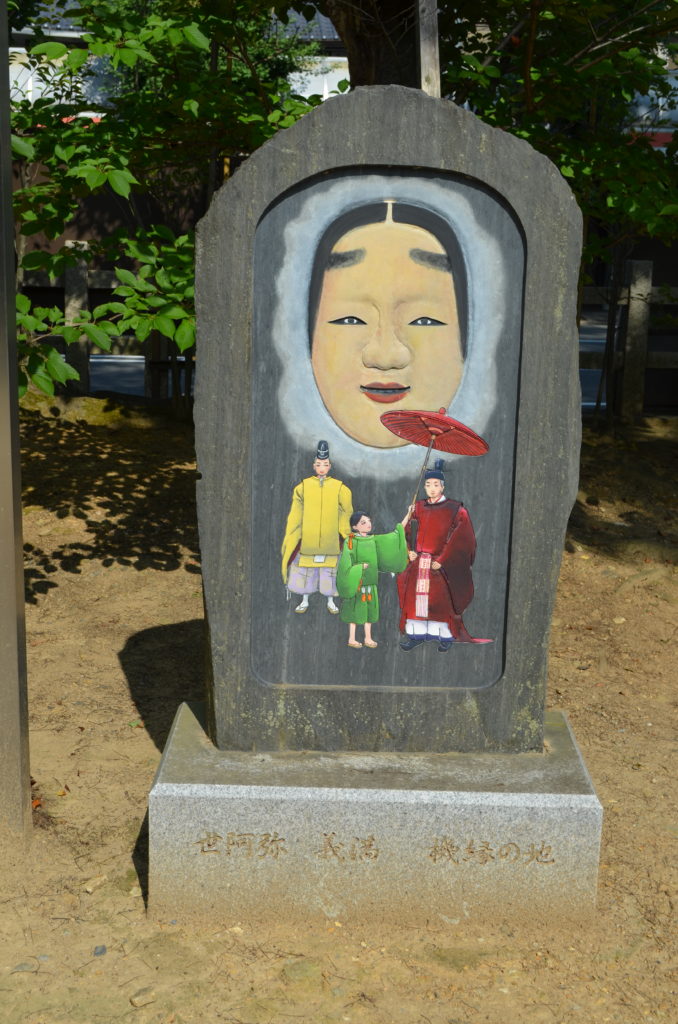 A rounded stone monument with a picture of a noh mask and three figures in traditional Japanese dress