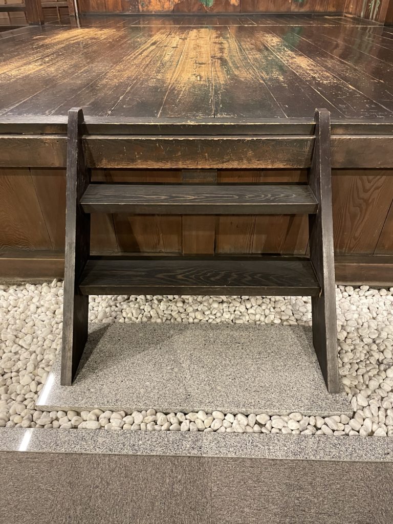 A wooden ladder with two steps descends from the front of the noh stage to a stone slab surrounded by white rocks.