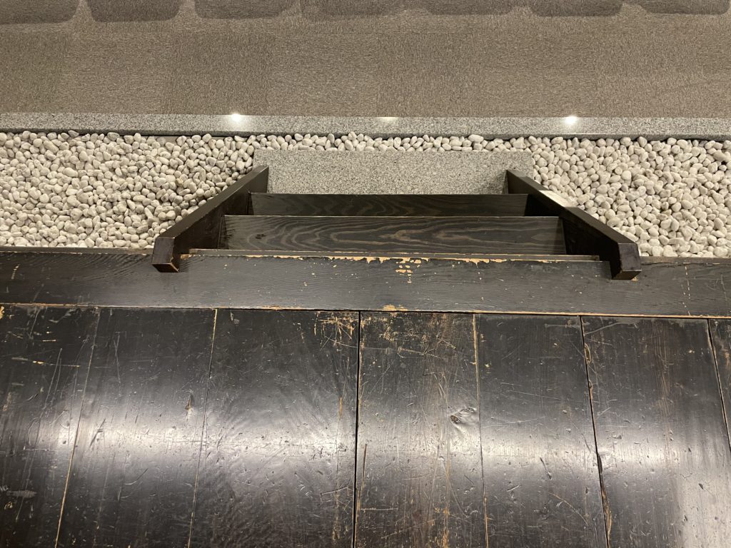 A bird's eye view of the short wooden staircase descending from the wooden noh stage to the moat of white rocks surrounding it.