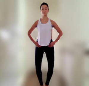 A woman in a white tanktop and black leggings stands against a blurred background with straight posture, elbows out and hands resting in front of her hips with her knees bent slightly outward.