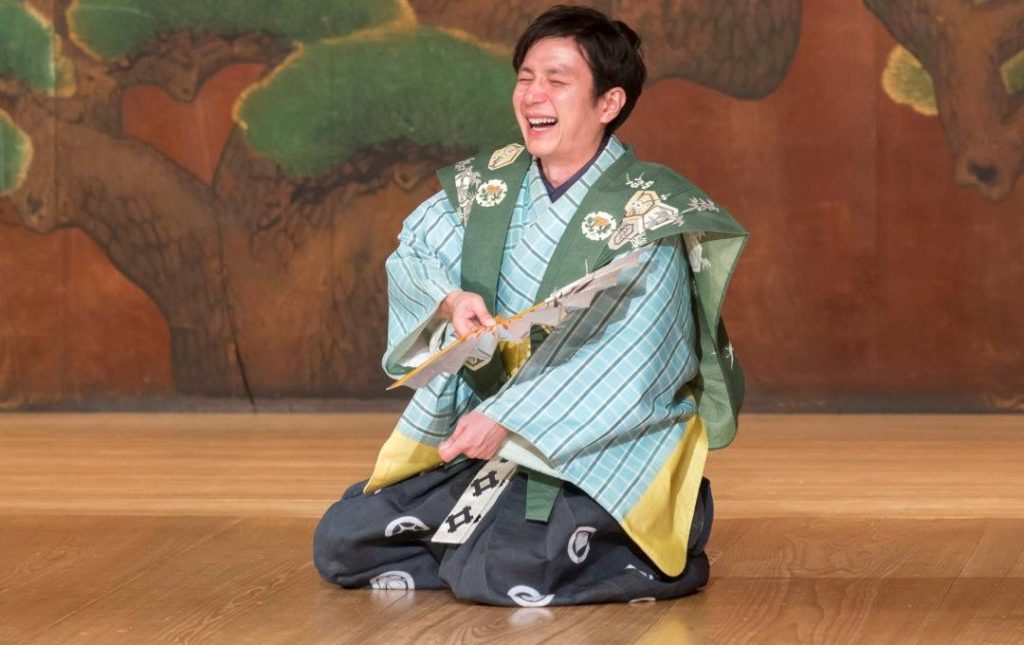 Shigeyama Sennojō III kneels on a traditional wooden stage in kyōgen costume laughing and holding an open fan