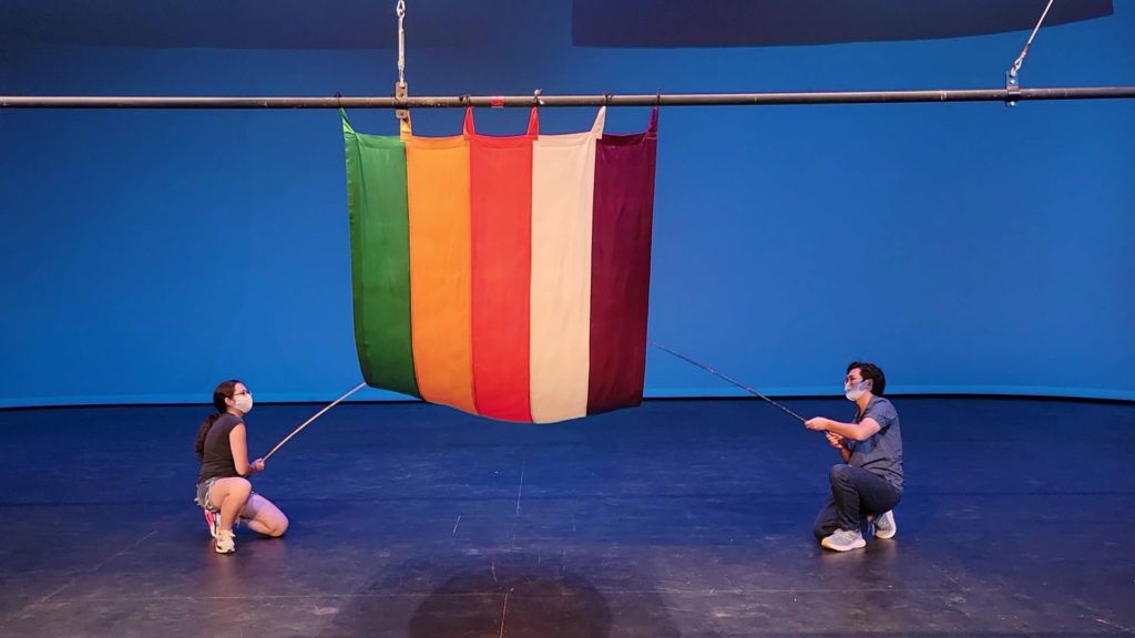 Two students use long, thin poles to pull up a green, yellow, red, white, and purple vertically striped curtain from the bottom.