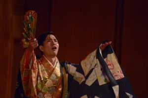 Shigeyama Sennojō III in an ornate kimono and outer robe holding a fanned looking upwards.