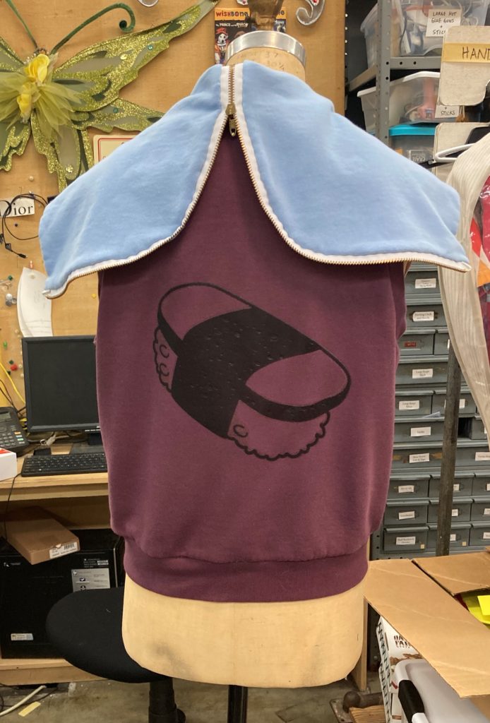 The backside of a purple vest with black line art of spam musubi and a zip open blue hood.