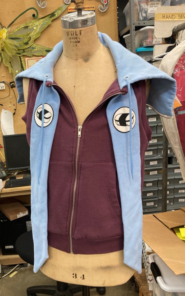 The front side of a purple vest with a blue zip open hood and blue strips on either side of the zipper running down the middle. The blue strips have a white and black spam musubi crest on them.