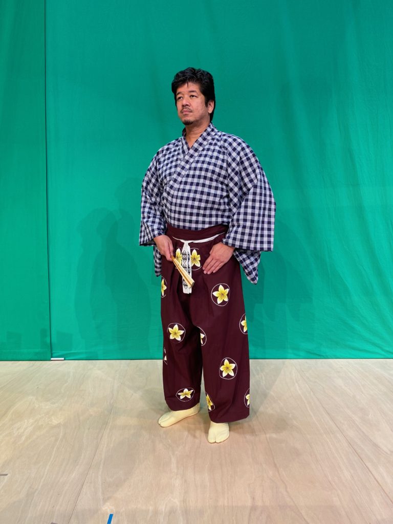 A man in a blue and white palaka kimono and purple pants with white and yellow plumeria crest holding a fan stands on a wooden floor in front of a green screen.