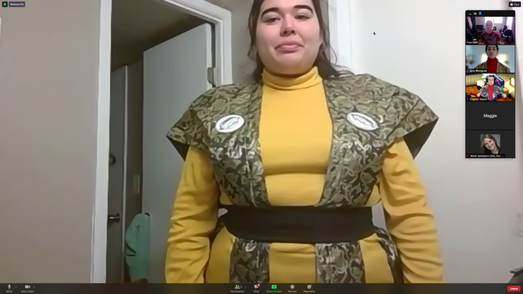 A screen shot of an actor wearing a yellow turtleneck with a green patterned kataginu over her shoulders and tied down around the waist with a black belt.