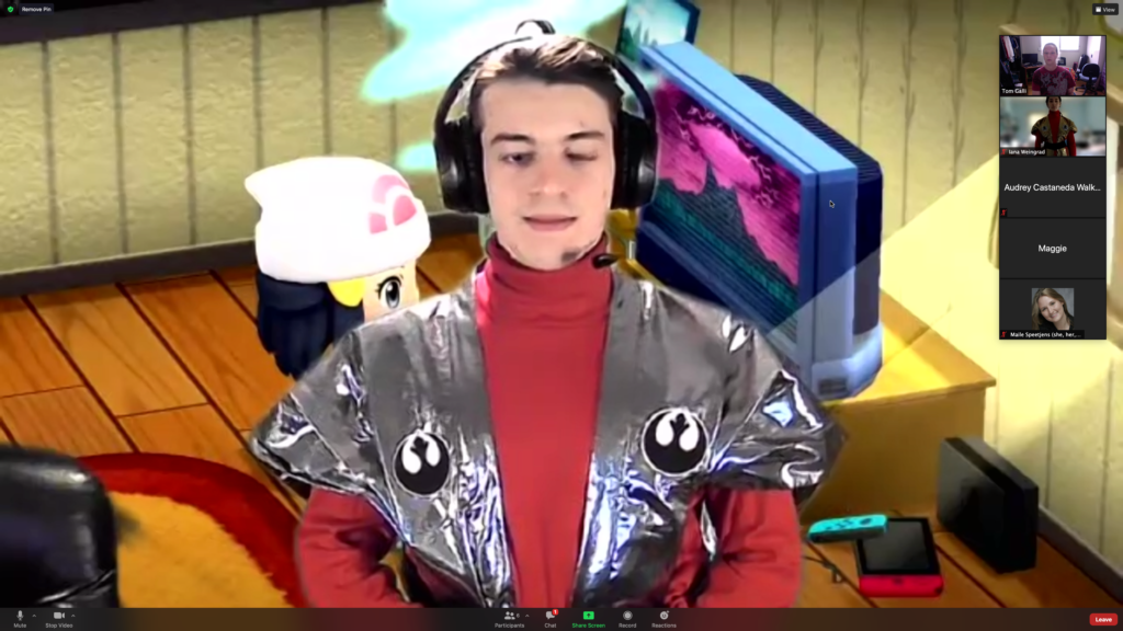 A screenshot of an actor standing in front of a pokemon virtual background wearing a red turtleneck, a silver vest with a black and white crest, and black headset.