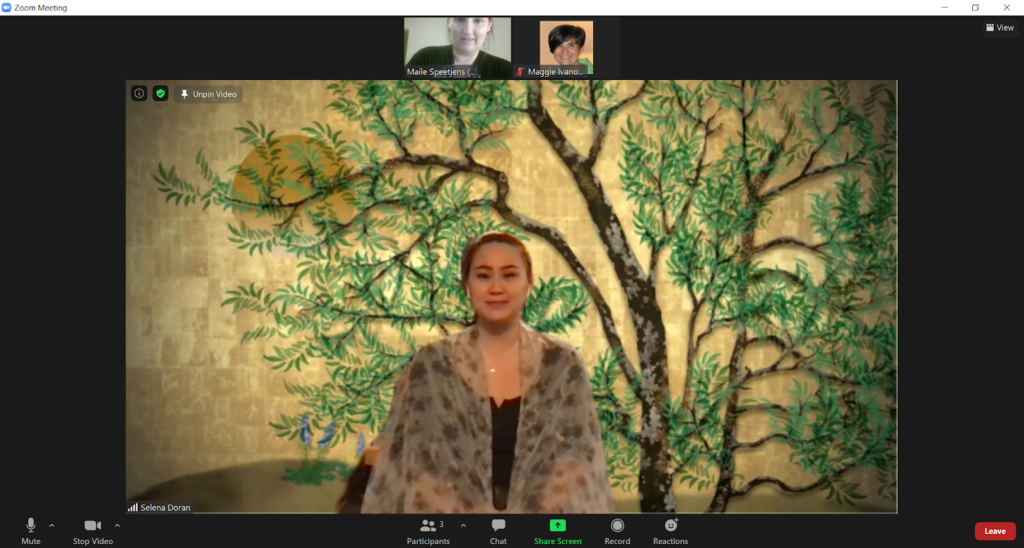 A screenshot of an actor sitting in front of a gold background with an illustration of a pecan tree wearing a black shirt with a translucent scarf draped over her shoulders.