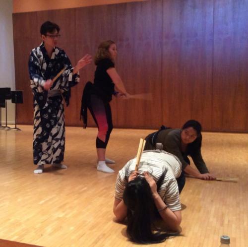 Shigeyama Sennojō III instructs a UHM student with a fan in hand while two more students duck on their hands and knees on the floor