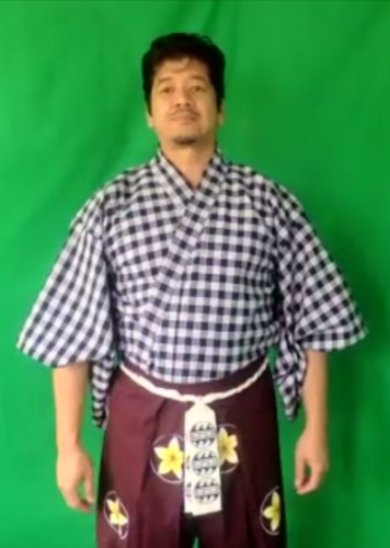 An actor stands in front of a green screen in a blue palaka kimono with a purple hakama with plumeria crests and a white koshi-obi.