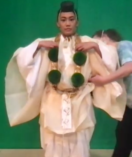 An actor stands in front of a green screen as a costume designer adjusts her white yamabushi kimono. She also wears green pompoms and a small, round, black hat.